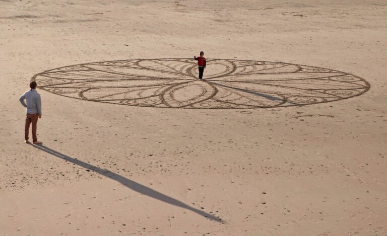 un bambino al centro di un grande mandala su una spiaggia con un adulto che lo guarda a distanza. I mandala di sabbia sono simbolo di impermanenza